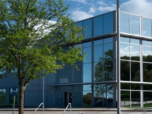 Iowa Department of Natural Resources office building with large glass windows, reflecting surrounding trees and sky. The building number 6200 is displayed near the entrance, with a parking lot and green landscape in the background