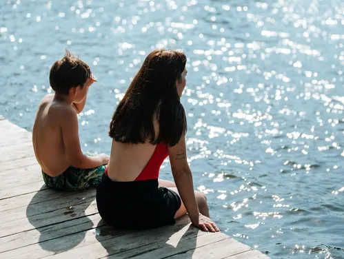 A young boy and a young woman sit on the edge of a dock on a lake