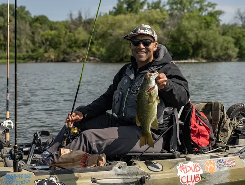 An angler in a kayak on a pond, wearing a life jacket, smiles as he holds up a nice smallmouth bass he caught.