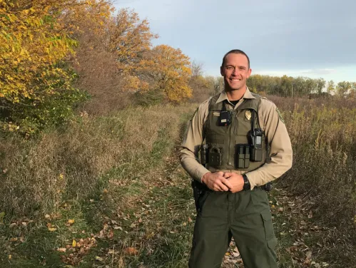 Conservation officer in full uniform standing along a trail near a treeline in the fall.