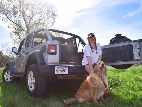 Woman sitting on the tailgate of her Jeep with her dog in a state park, showing off her Iowa natural resources license plate.