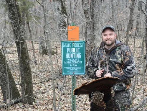 Turkey hunter with a harvested turkey standing next to a public hunting wildlife management area sign.