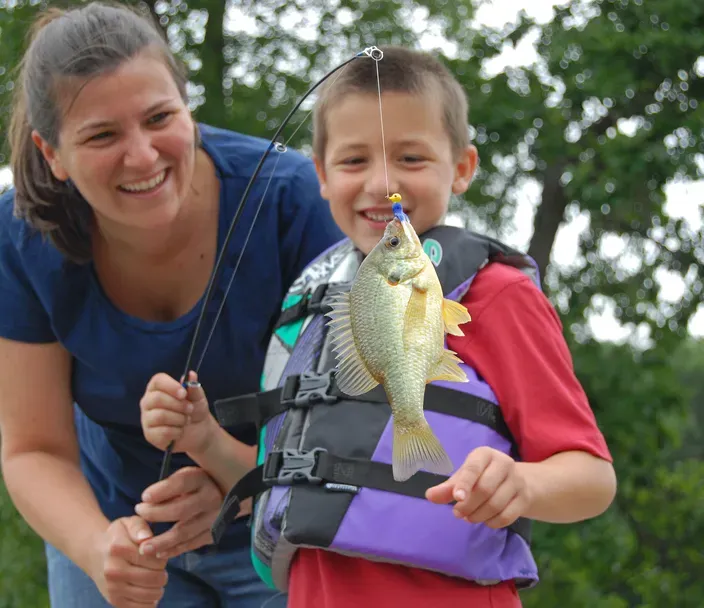 A mother smiles as she and her son, wearing a life jacket, look at the fish on the end of his line while standing a dock in a park.