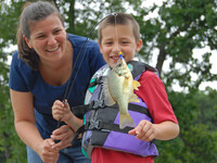 A mother smiles as she and her son, wearing a life jacket, look at the fish on the end of his line while standing a dock in a park.