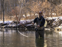 A man with a beard in long waders fishes for trout in a stream