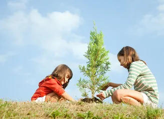 Two kids sitting on the ground with lots of blue sky behind them as they plant a tree.