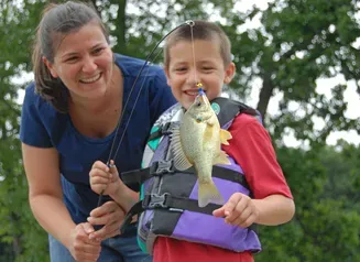 A mother smiles as she and her son, wearing a life jacket, look at the fish on the end of his line while standing a dock in a park.