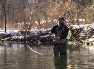 A man with a beard in long waders fishes for trout in a stream