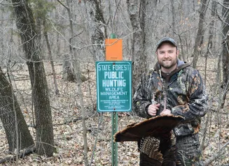 Turkey hunter with a harvested turkey standing next to a public hunting wildlife management area sign.